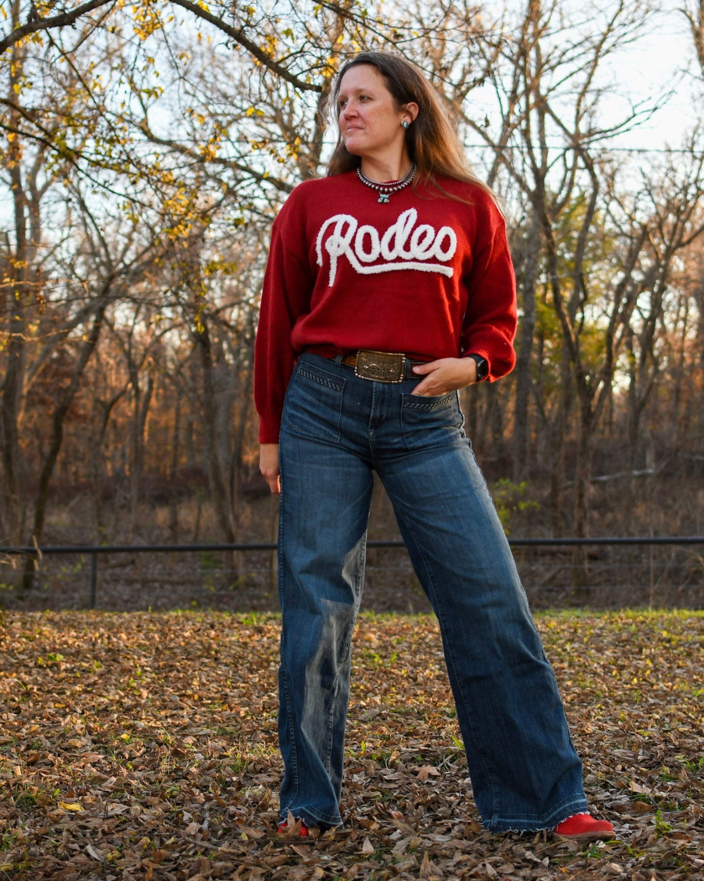 Woman standing in trees wearing a rodeo sweater and jeans 