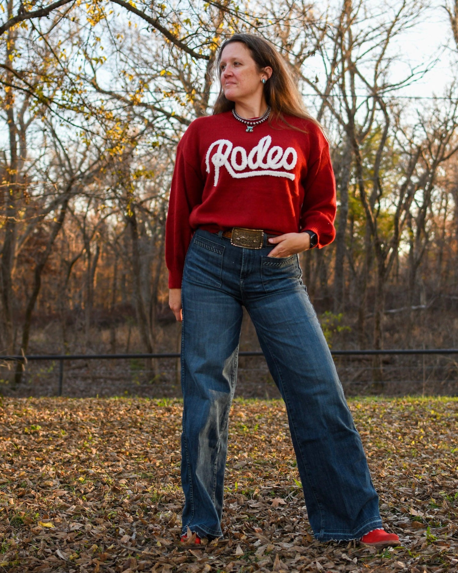 Woman standing in trees wearing a rodeo sweater and jeans 