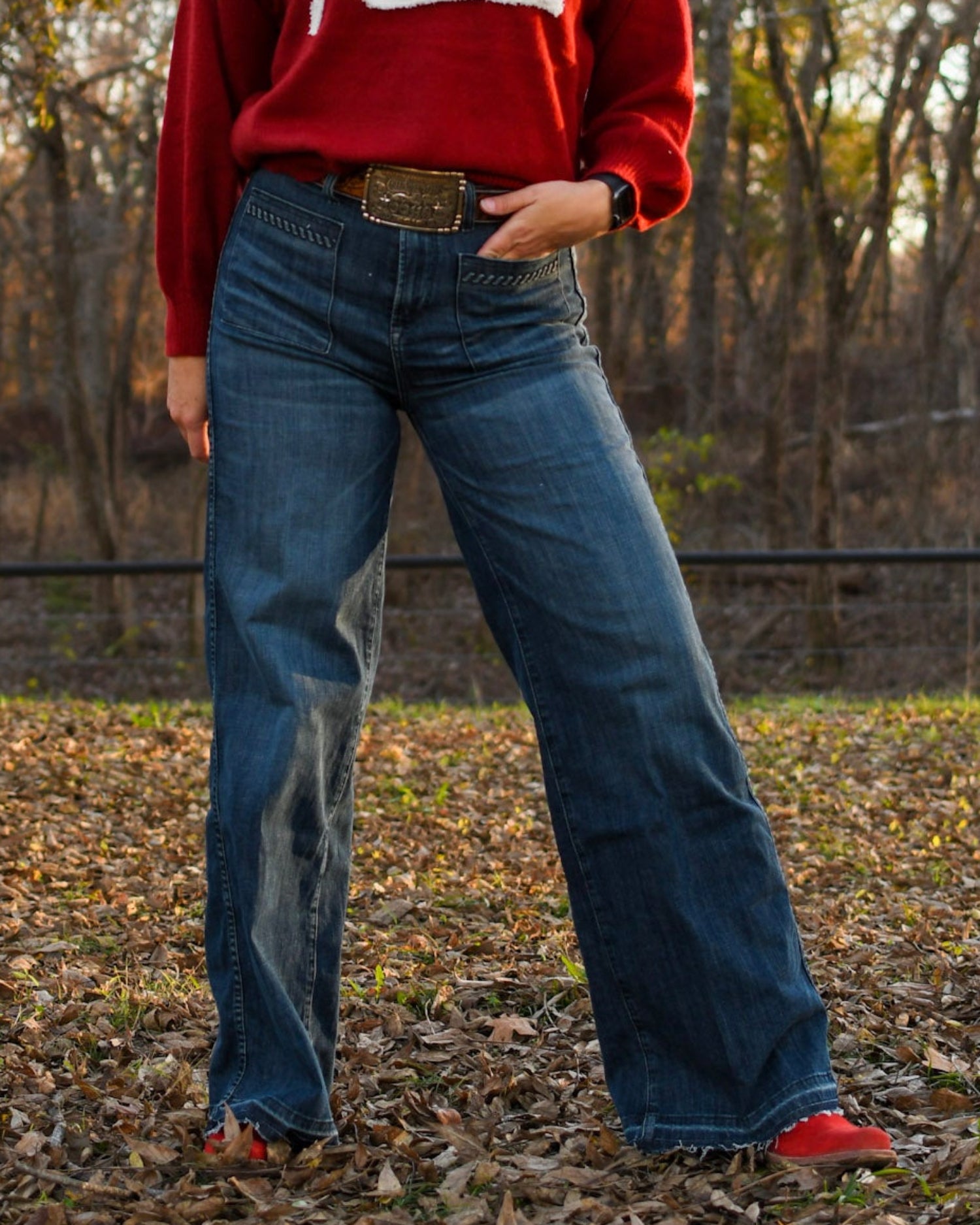 woman standing in trees wearing jeans and a sweater