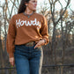 Model wearing Howdy sweater in front of fence with trees