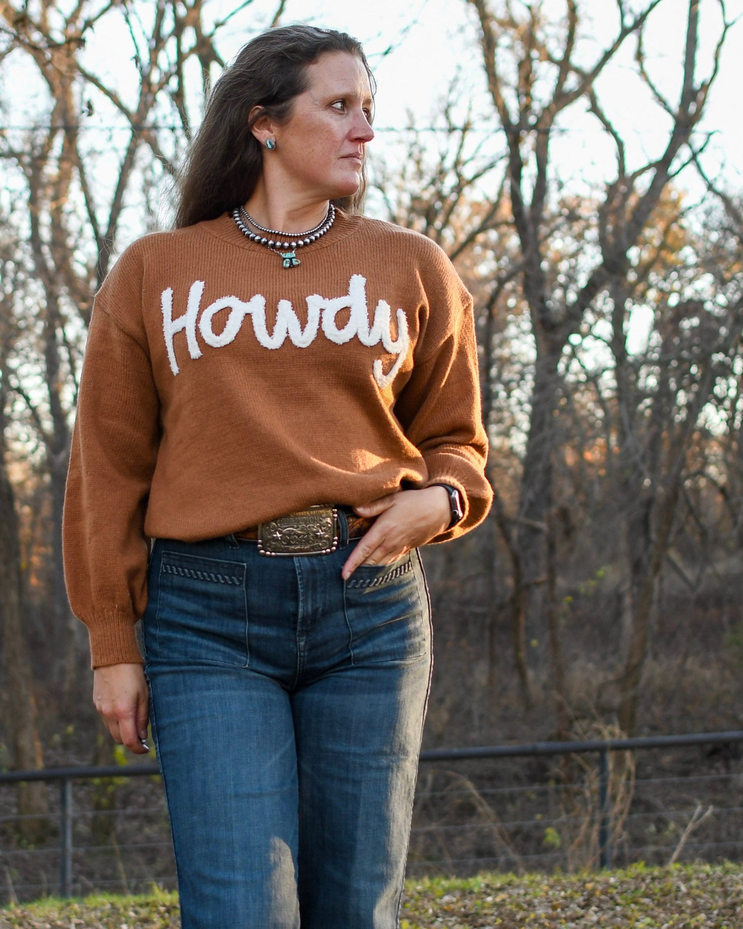 Model wearing Howdy sweater in front of fence with trees