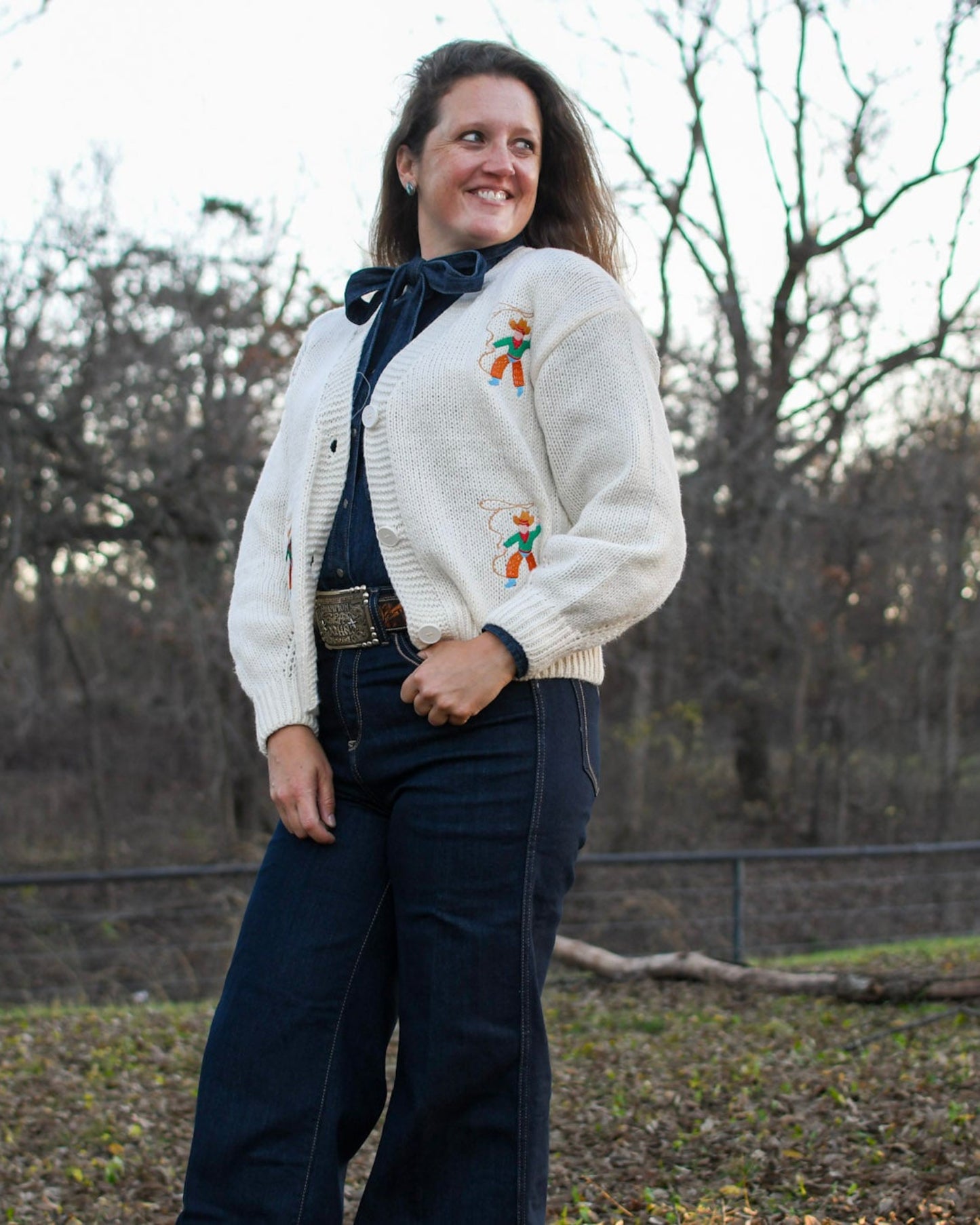 woman standing in front of trees wearing jeans and a cardigan sweater