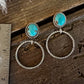 A pair of silver-toned dangle earrings with oval turquoise-blue stones and twisted hoop drops, displayed on a rustic wooden surface next to a metal ruler for scale.