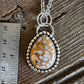 A silver beaded pendant necklace with a teardrop-shaped orange and white patterned stone, displayed next to a metal ruler on a rustic wooden surface.