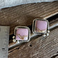 A pair of square stud earrings with pink stone in a silver rope-style bezel setting, placed on a wooden surface next to a metal ruler for scale.