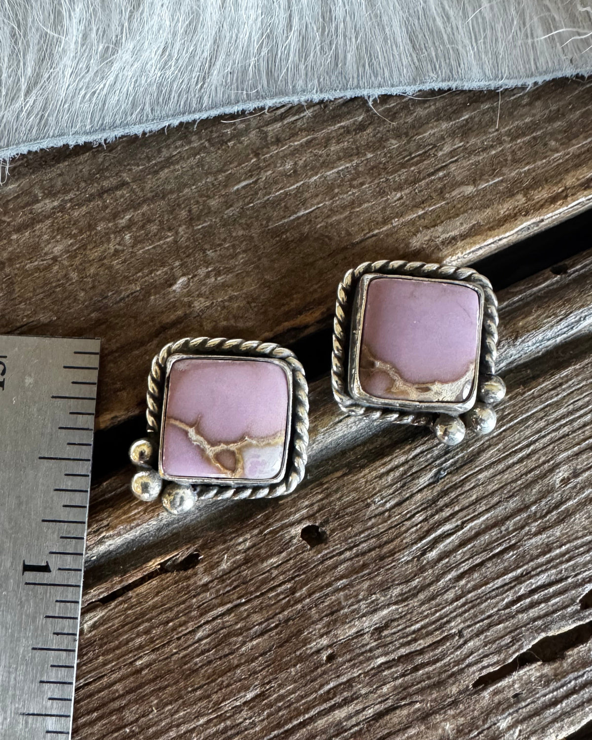 A pair of square stud earrings with pink stone in a silver rope-style bezel setting, placed on a wooden surface next to a metal ruler for scale.