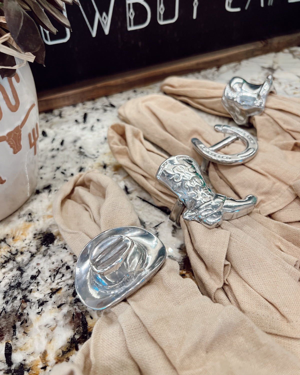 Silver cowboy boot and hat napkin rings on beige fabric with a 'Cowboy Cafe' sign in the background.