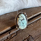 An oval turquoise ring with a silver-toned twisted border, resting on a rustic wooden surface with white fur in the background.