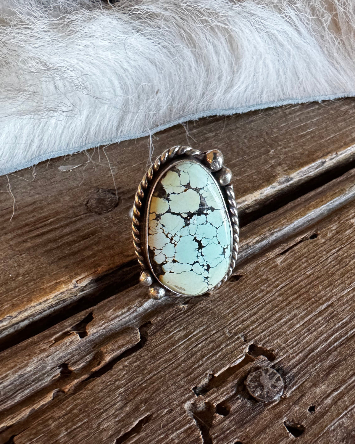 An oval turquoise ring with a silver-toned twisted border, resting on a rustic wooden surface with white fur in the background.