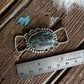 Silver pendant with a turquoise stone on a wooden surface with a ruler for scale.