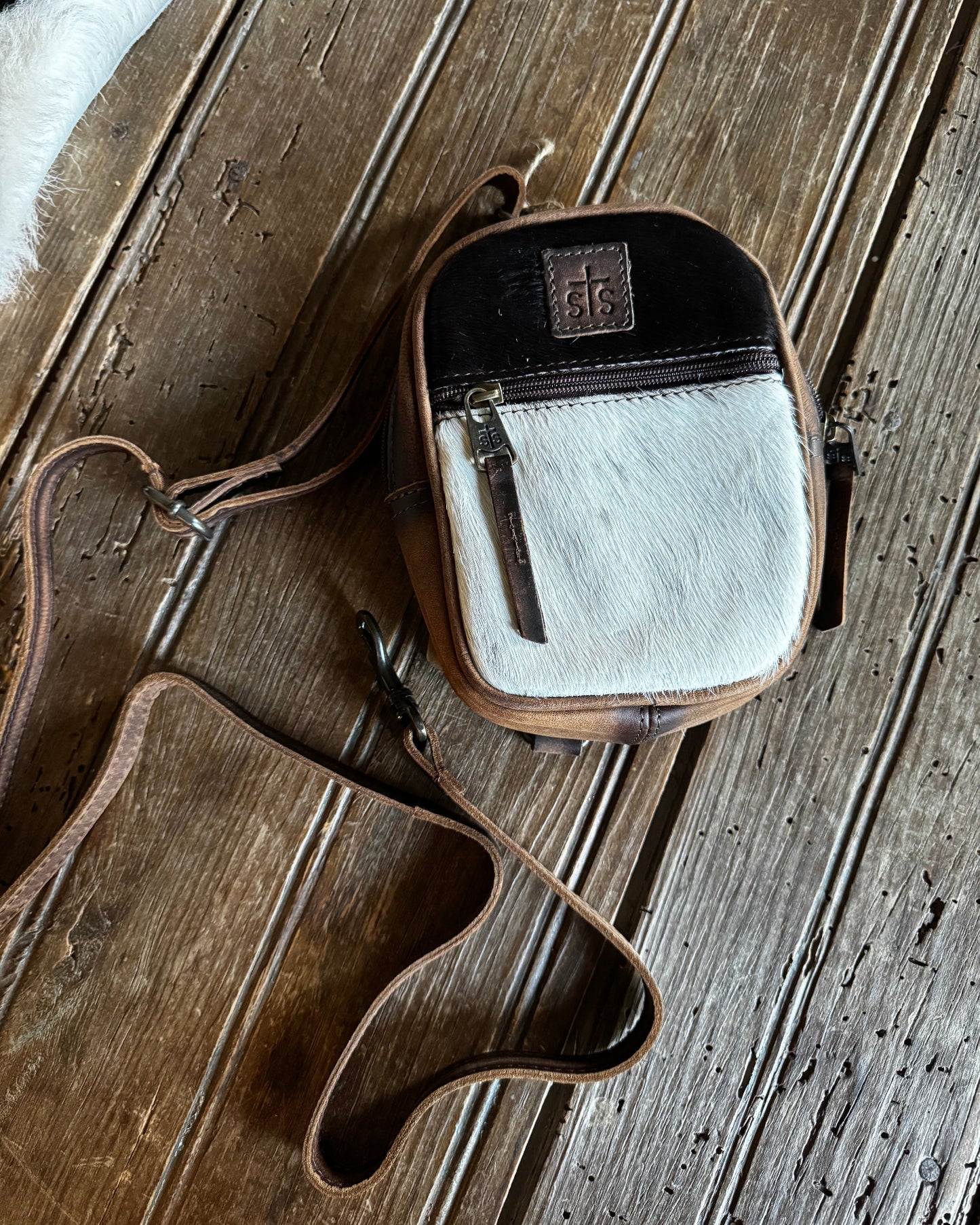 A small crossbody bag with a brown leather body, a black top panel, and a white cowhide front panel, shown on a rustic wooden plank surface. The bag has a long brown leather strap and a small rectangular logo patch on the front.