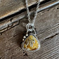 A silver necklace with a triangular pendant featuring a textured yellow and brown stone, displayed on a rustic, weathered wooden surface.