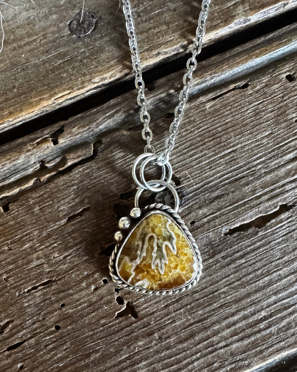 A silver necklace with a triangular pendant featuring a textured yellow and brown stone, displayed on a rustic, weathered wooden surface.