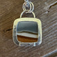 A silver pendant necklace with a square-shaped bezel containing a polished band of brown and black agate, resting on a rustic dark wood surface.