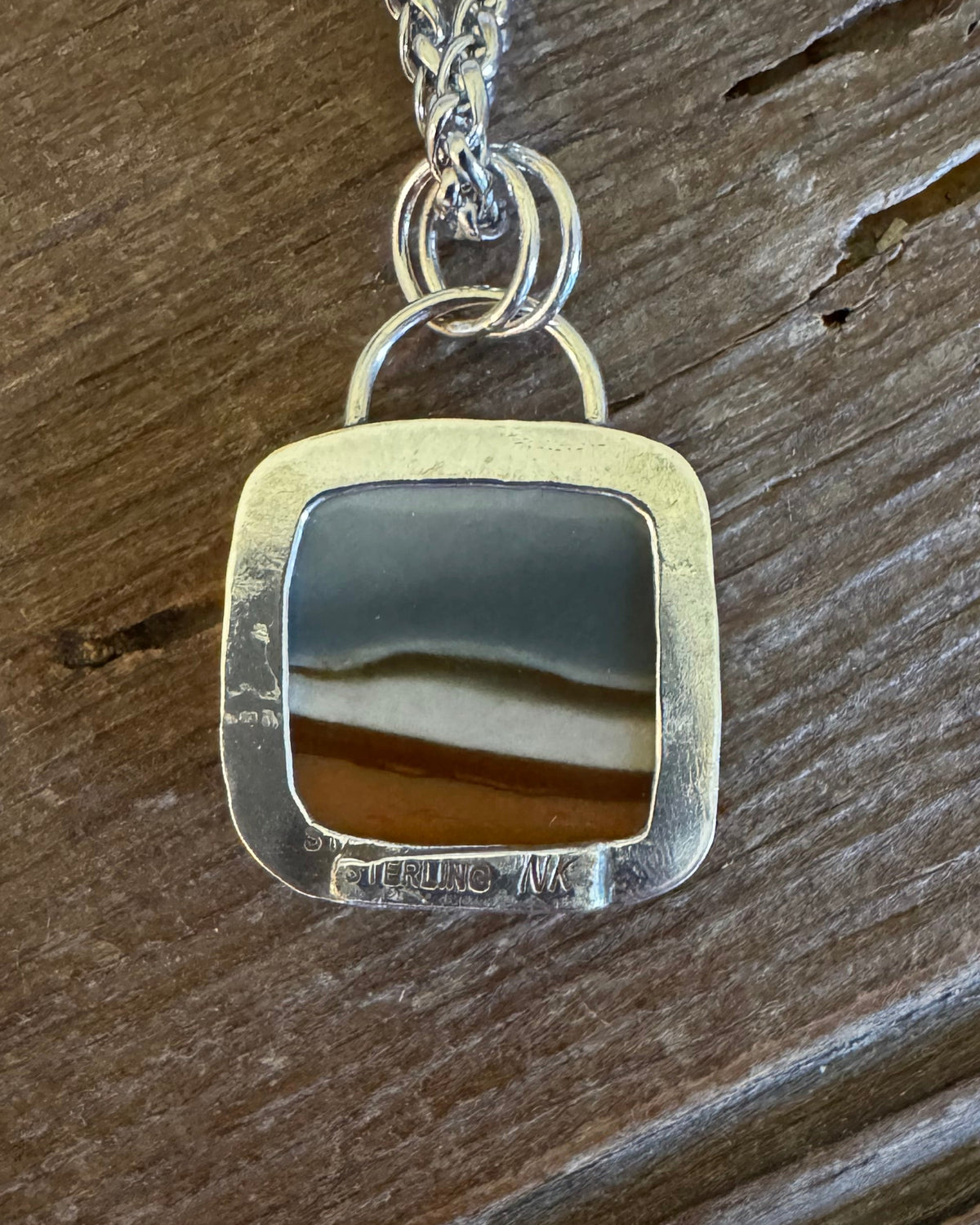 A silver pendant necklace with a square-shaped bezel containing a polished band of brown and black agate, resting on a rustic dark wood surface.
