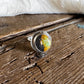 A silver ring featuring a round cabochon gemstone with a yellow and black marbled pattern, resting on a rustic, weathered wooden surface with a white and black cowhide pelt in the background.