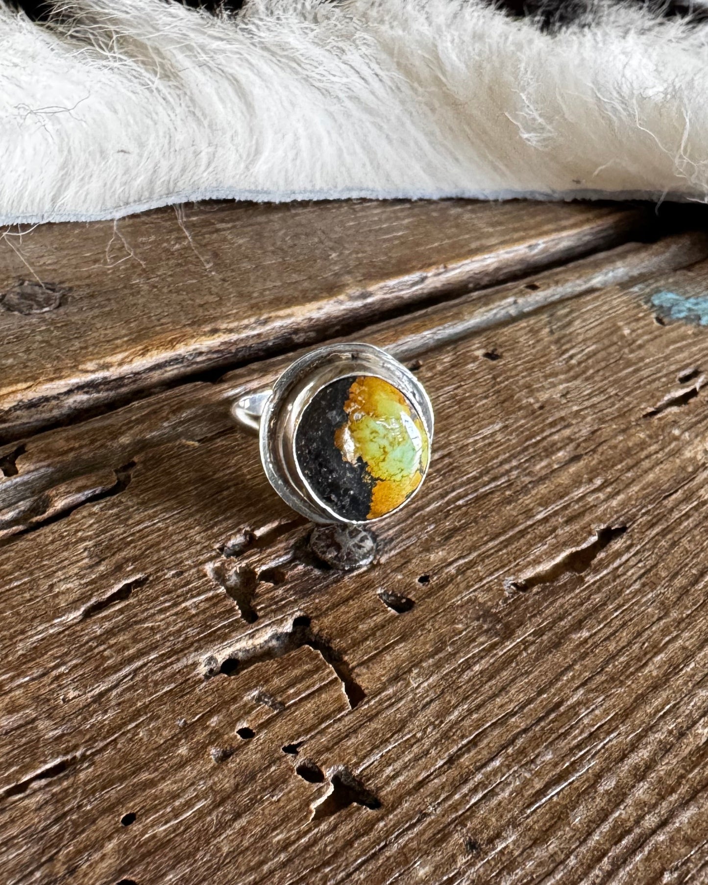 A silver ring featuring a round cabochon gemstone with a yellow and black marbled pattern, resting on a rustic, weathered wooden surface with a white and black cowhide pelt in the background.