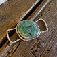 A silver-toned slide with a green stone, resting on a rustic dark wood surface with a white fur trim visible at the top.