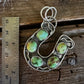 A sterling silver horseshoe-shaped pendant featuring five green stone inlays, displayed next to a metal ruler on a rustic wooden surface.