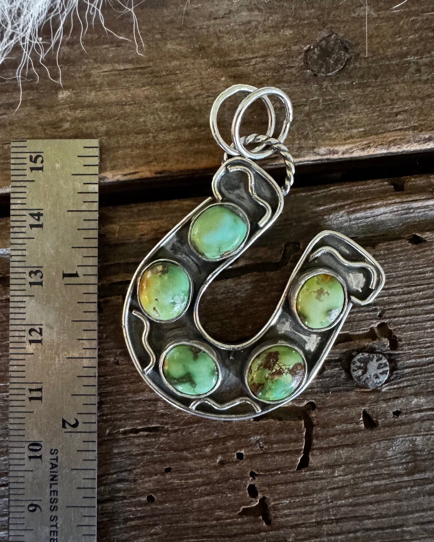 A sterling silver horseshoe-shaped pendant featuring five green stone inlays, displayed next to a metal ruler on a rustic wooden surface.