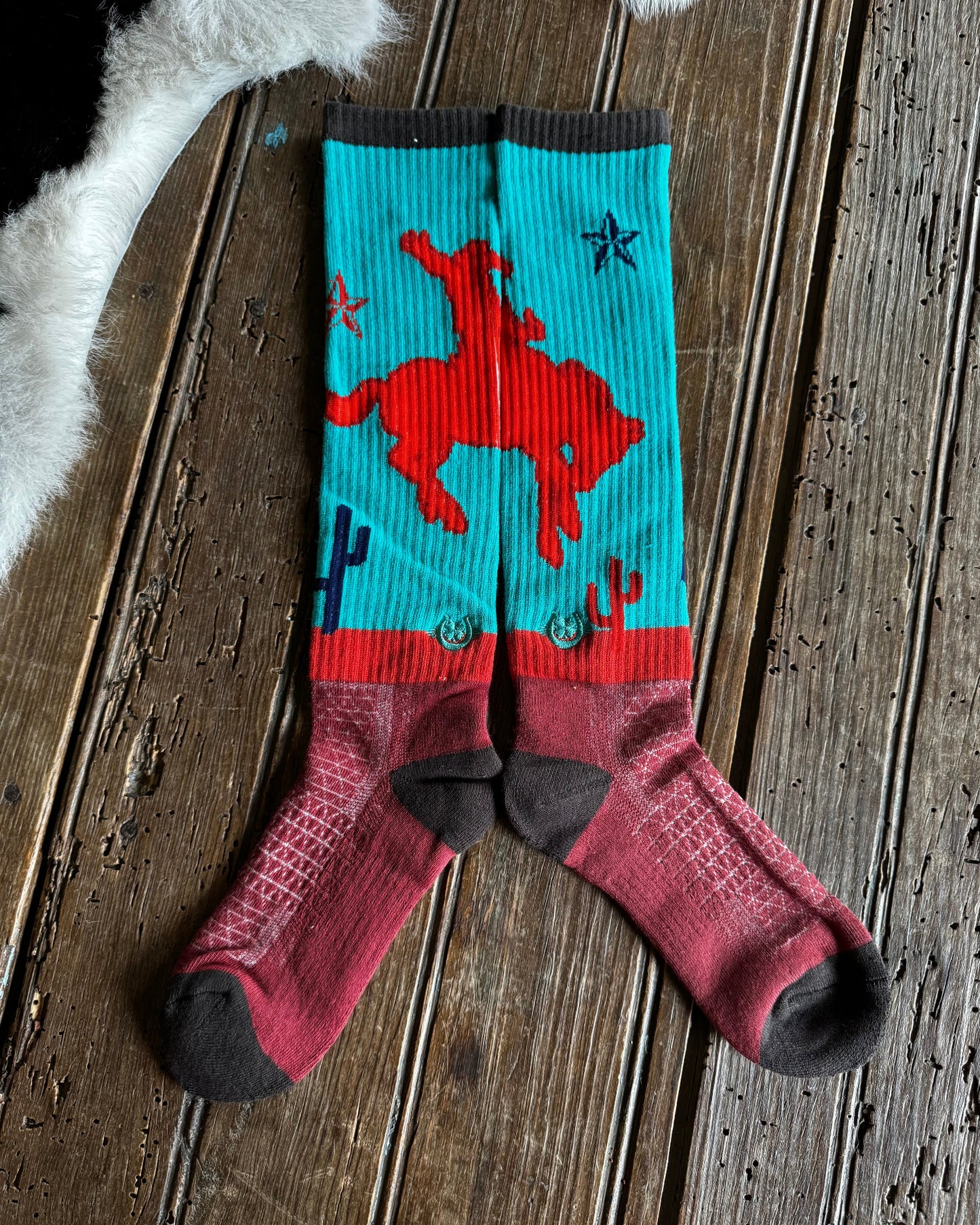 A pair of knee-high socks laid flat on a rustic wooden surface. The socks feature a turquoise upper section with a red cowboy riding a bucking horse, dark gray cuffs and heels, and a dark red lower section with textured knit patterns.