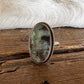 A silver-toned ring with an oval green and brown stone, standing upright on a rustic wooden surface in front of a black and white fur rug.