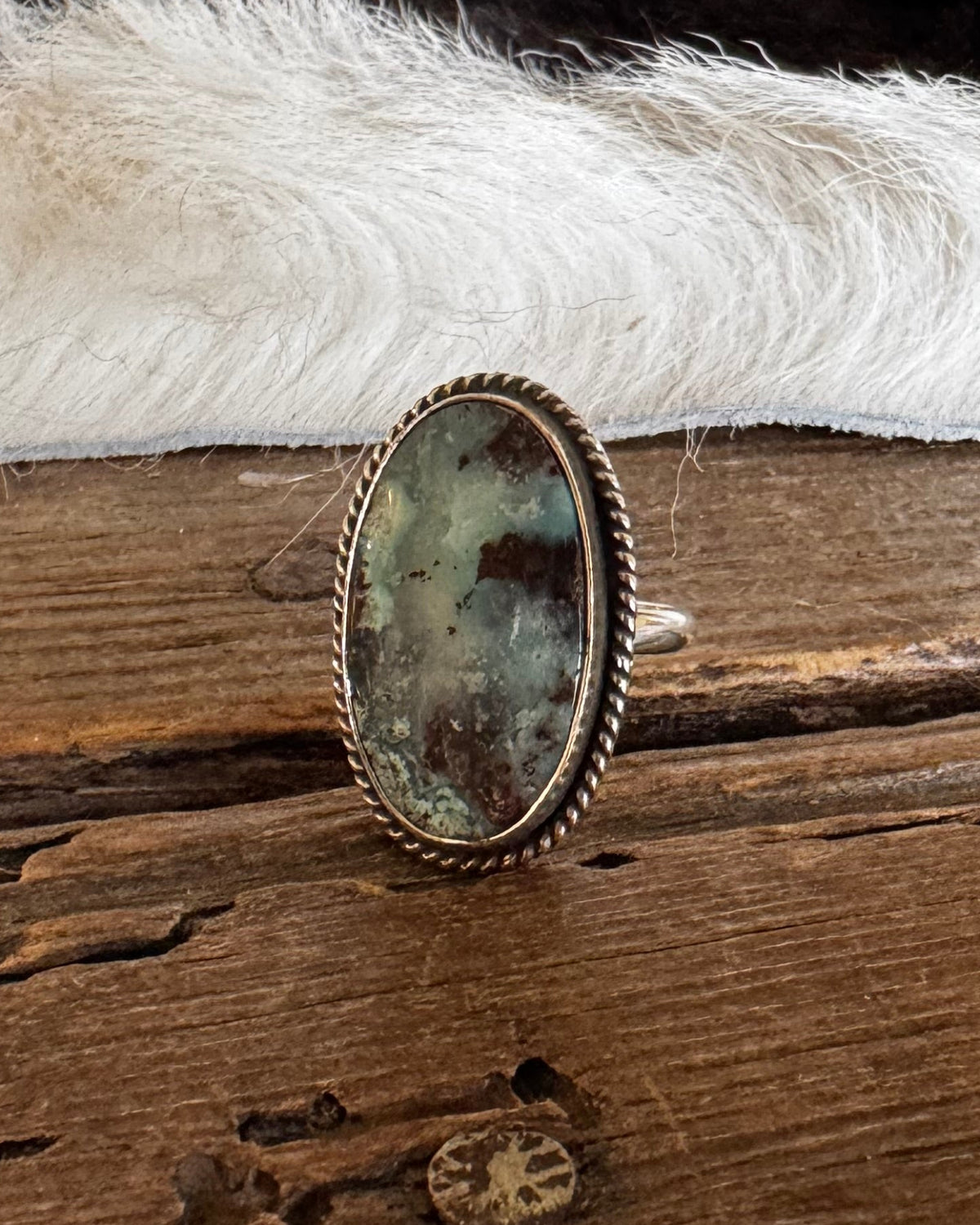 A silver-toned ring with an oval green and brown stone, standing upright on a rustic wooden surface in front of a black and white fur rug.
