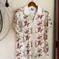 A short-sleeve collared button-up shirt with a western cowboy print, hanging on a wooden hanger from a black wall hook against a beige wall.