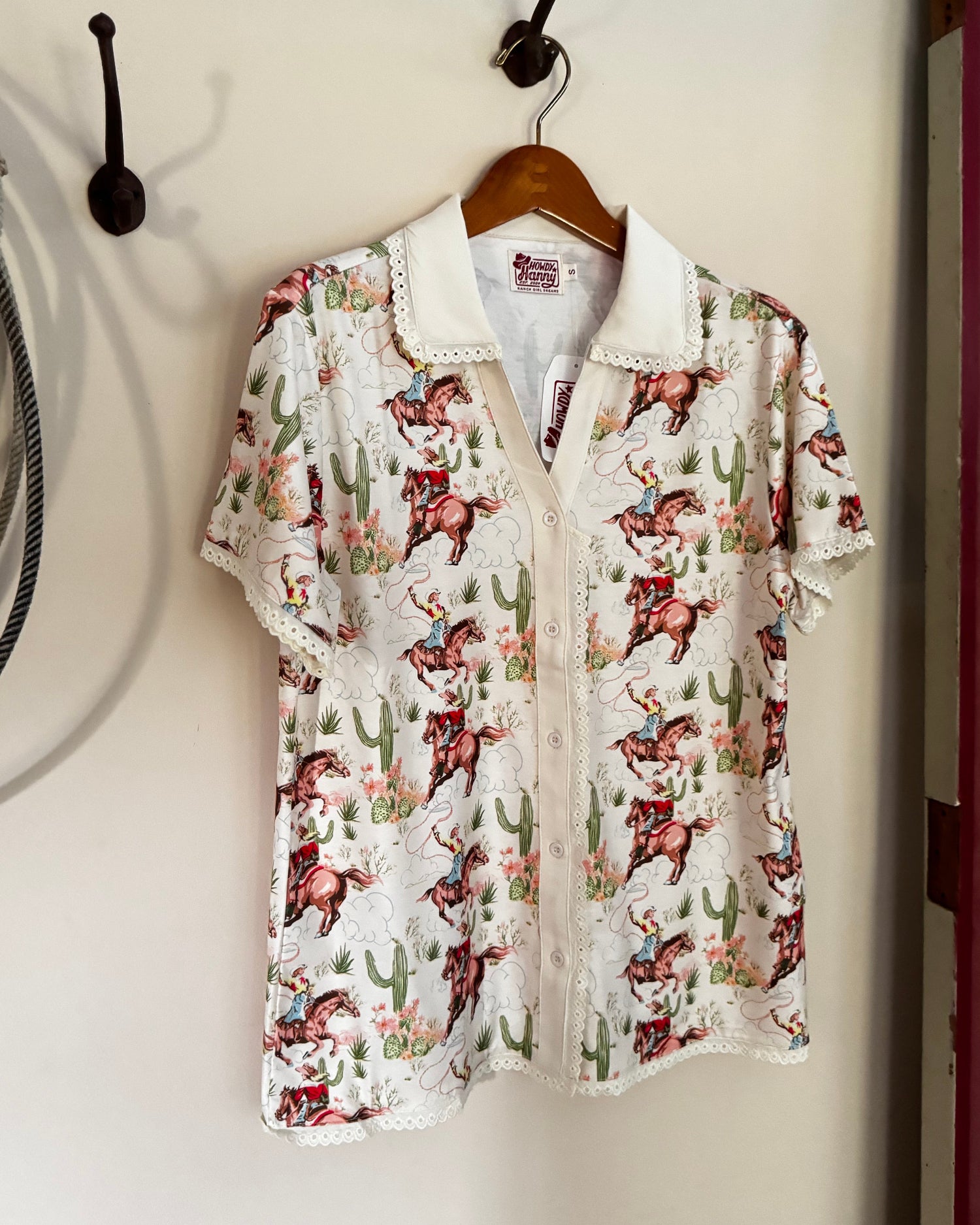 A short-sleeve collared button-up shirt with a western cowboy print, hanging on a wooden hanger from a black wall hook against a beige wall.