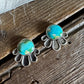 A pair of silver-toned stud earrings with oval turquoise-blue stones and petal-like silver settings, resting on a rustic wooden surface with a white faux-fur textile at the top.