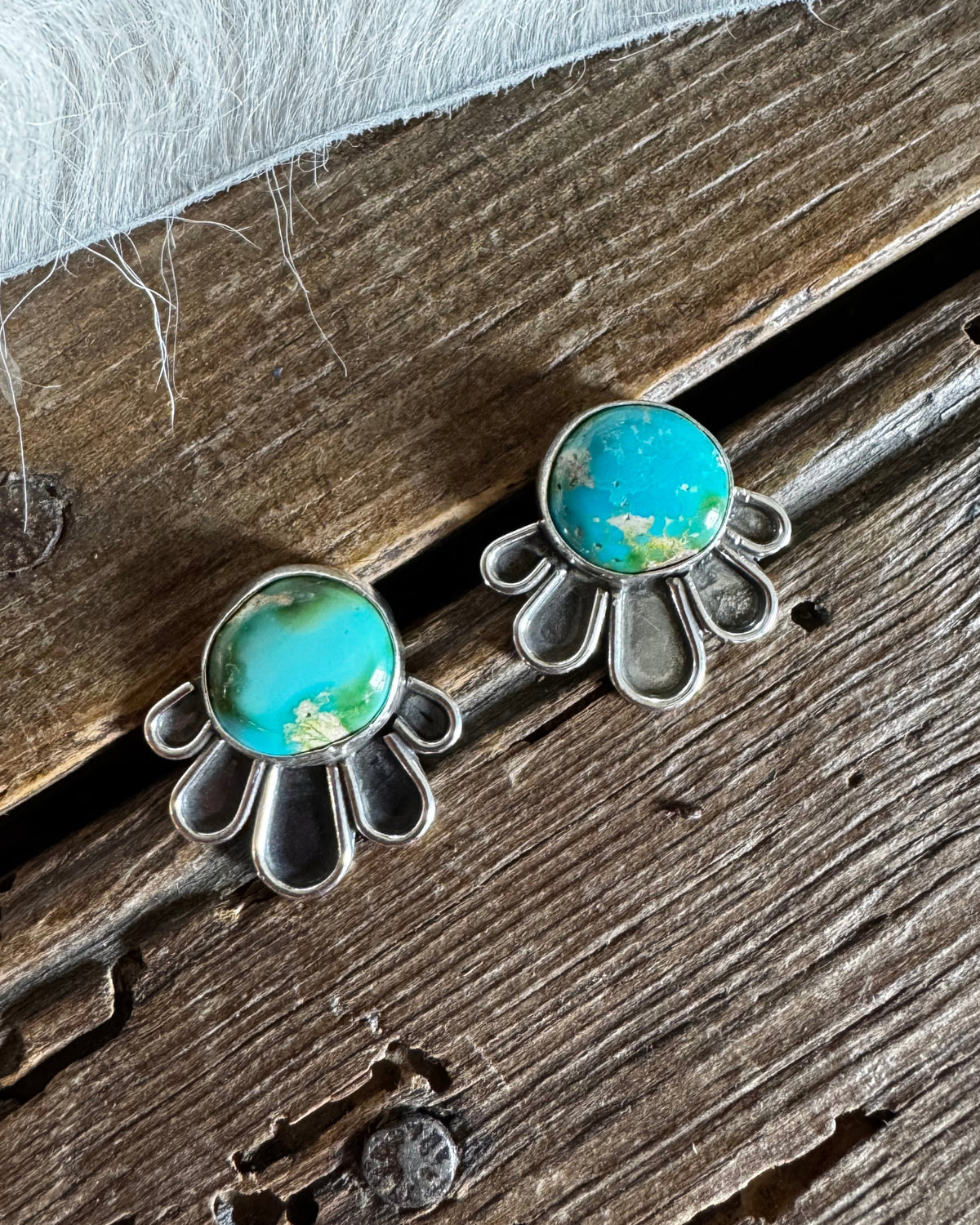 A pair of silver-toned stud earrings with oval turquoise-blue stones and petal-like silver settings, resting on a rustic wooden surface with a white faux-fur textile at the top.