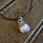 A teardrop-shaped purple and white marbled gemstone pendant set in a silver bezel, hanging from a black cord necklace on a rustic dark wood background.