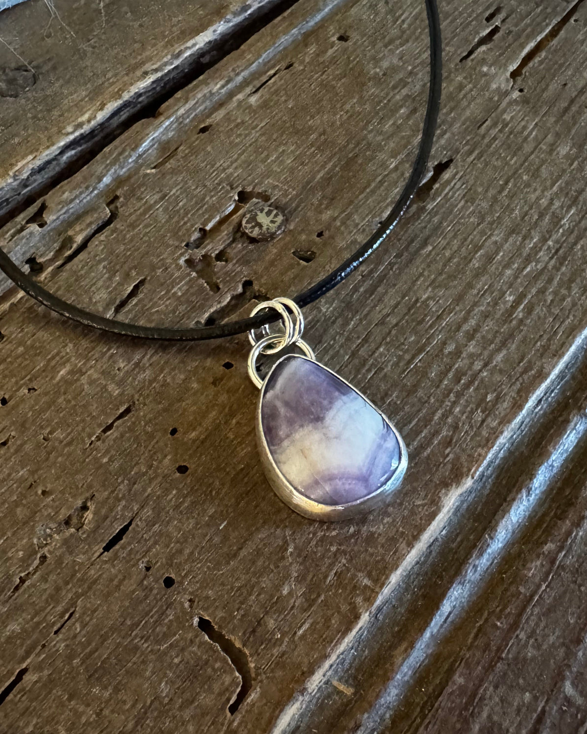 A teardrop-shaped purple and white marbled gemstone pendant set in a silver bezel, hanging from a black cord necklace on a rustic dark wood background.