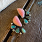 A pair of handmade turquoise and coral pink stud earrings with silver-toned metal settings, displayed on a rustic dark wood surface next to a white fur trim.