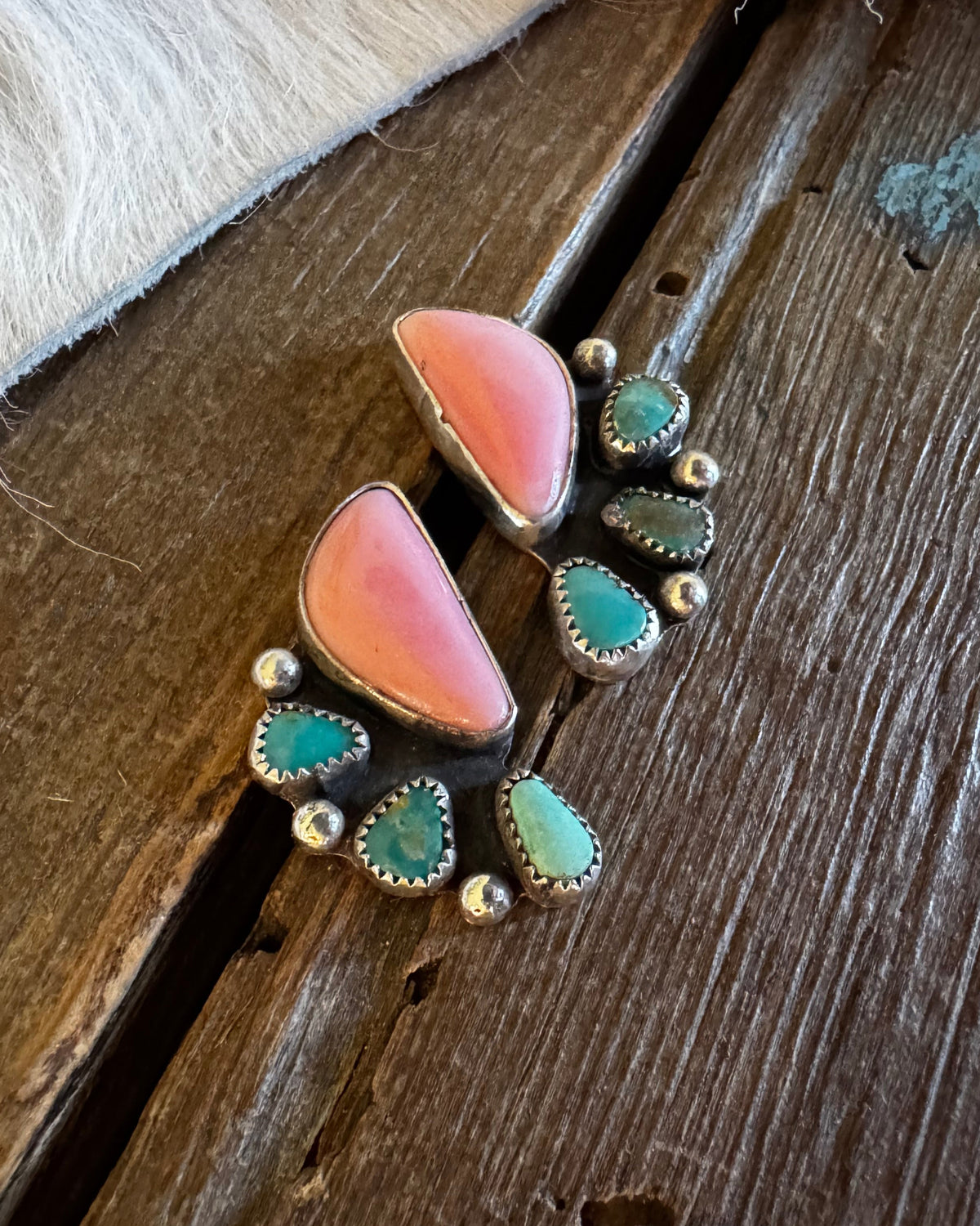 A pair of handmade turquoise and coral pink stud earrings with silver-toned metal settings, displayed on a rustic dark wood surface next to a white fur trim.