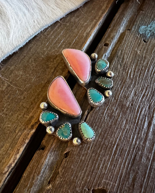 A pair of handmade turquoise and coral pink stud earrings with silver-toned metal settings, displayed on a rustic dark wood surface next to a white fur trim.