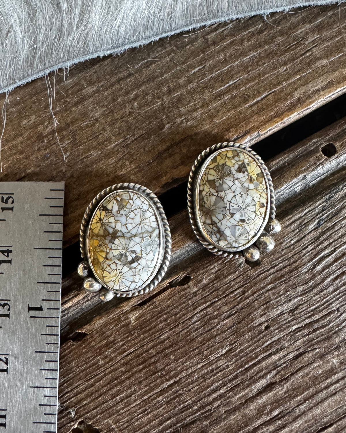 A pair of oval stud earrings with textured cream and gold-toned stone centers in silver-toned bezel settings, displayed on a rustic wooden surface next to a metal ruler for scale.