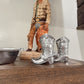 Silver cowboy boots on a wooden surface with a figurine in the background.