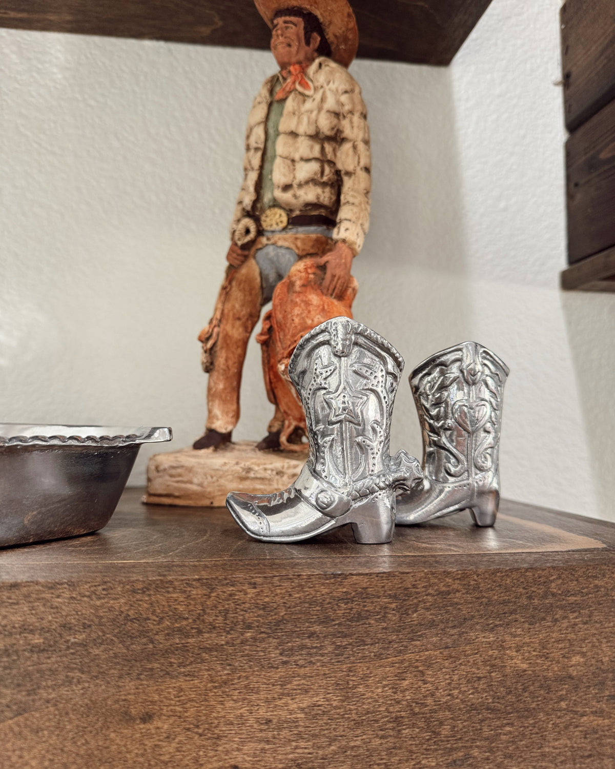 Silver cowboy boots on a wooden surface with a figurine in the background.