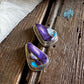 A pair of triangular stud earrings with purple and turquoise inlay set in silver, resting on a rustic wooden surface next to a white faux fur trim.