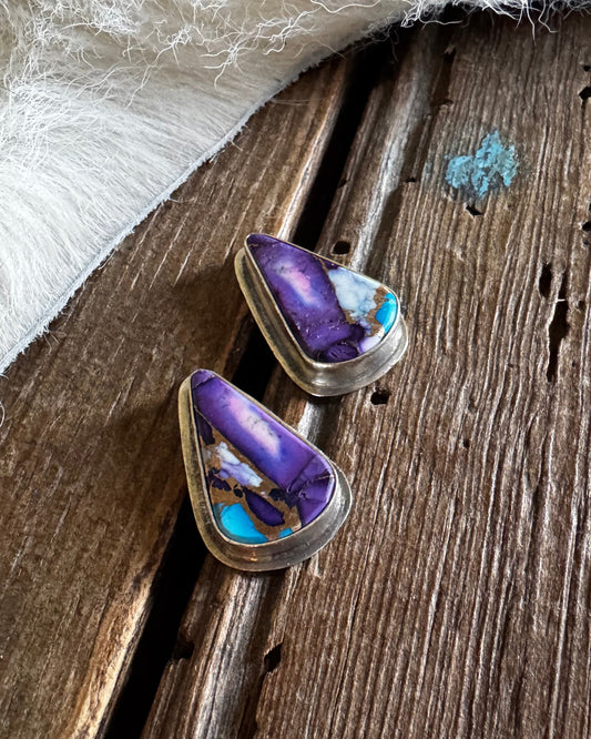A pair of triangular stud earrings with purple and turquoise inlay set in silver, resting on a rustic wooden surface next to a white faux fur trim.
