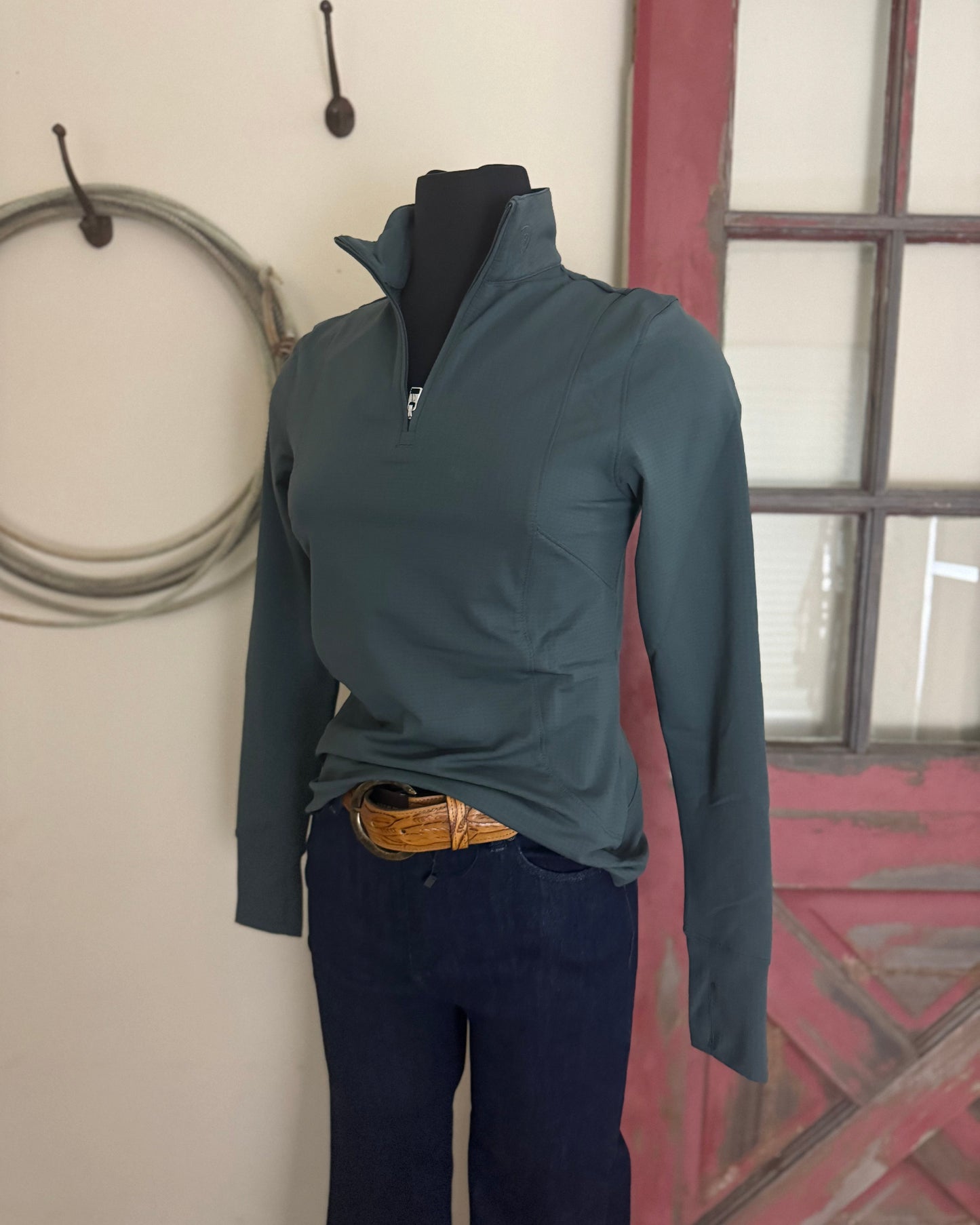 Mannequin wearing a teal long-sleeve shirt and dark pants against a wall with a window.