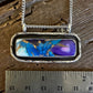 A silver-toned rectangular pendant necklace with a marbled blue, purple, and gold inlay, displayed on a rustic wooden surface next to a metal ruler for scale.