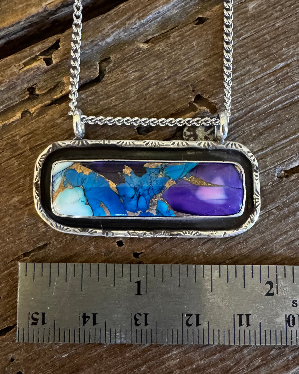 A silver-toned rectangular pendant necklace with a marbled blue, purple, and gold inlay, displayed on a rustic wooden surface next to a metal ruler for scale.