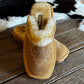 A pair of tan suede slip-on slippers with white embroidered western-style floral details and a white faux-fur trim, displayed on a rustic wooden table with a cowhide rug in the background.
