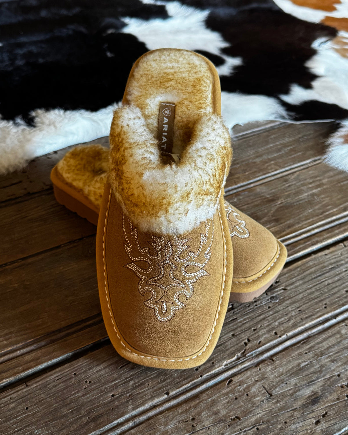 A pair of tan suede slip-on slippers with white embroidered western-style floral details and a white faux-fur trim, displayed on a rustic wooden table with a cowhide rug in the background.