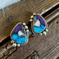 A pair of silver-toned teardrop-shaped stud earrings featuring a marbled turquoise, purple, and white stone inlay, displayed on a rustic dark wood surface next to a white faux fur texture.