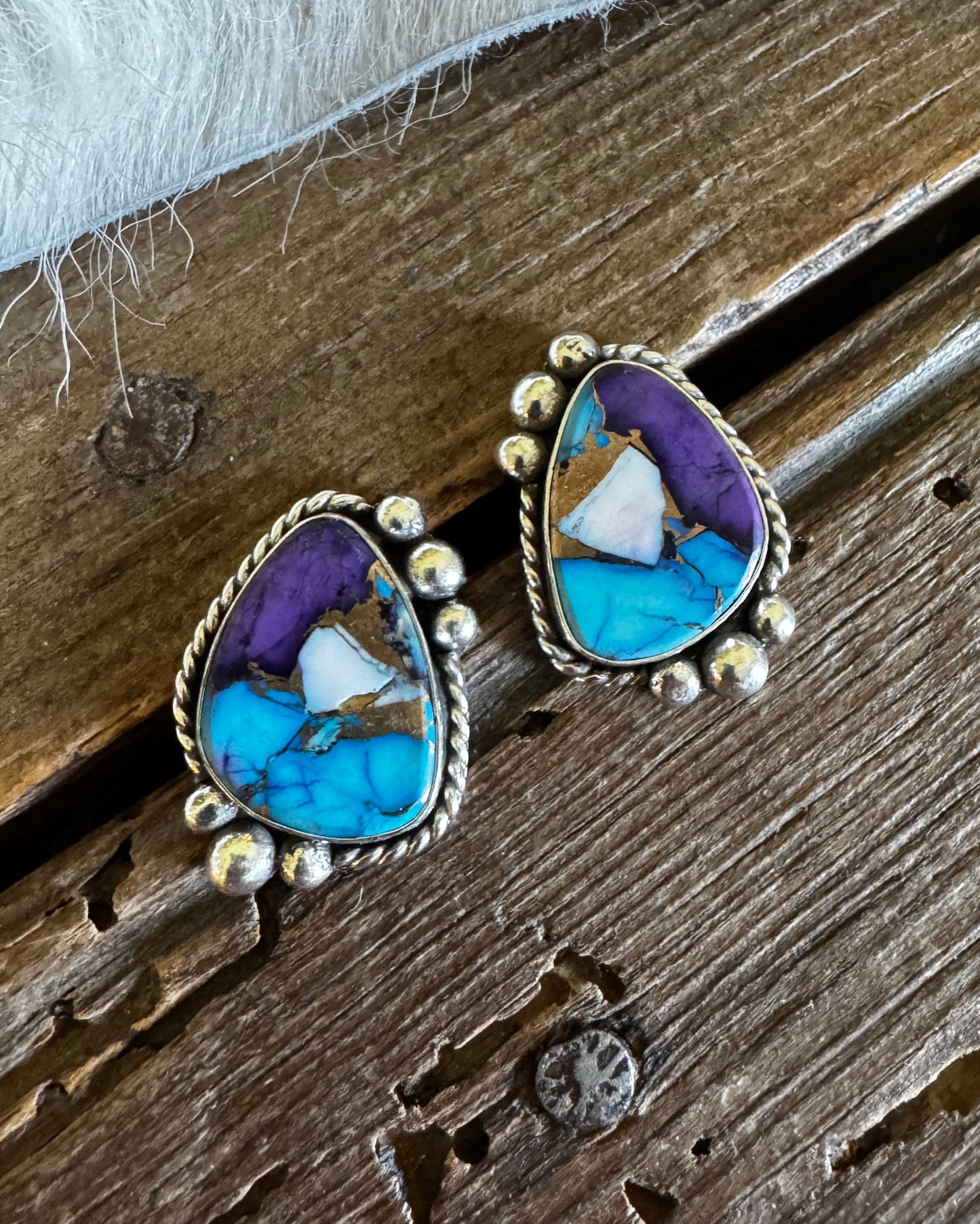 A pair of silver-toned teardrop-shaped stud earrings featuring a marbled turquoise, purple, and white stone inlay, displayed on a rustic dark wood surface next to a white faux fur texture.
