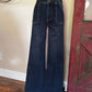 Dark blue wide-leg denim jeans displayed on a black mannequin torso in an indoor room with a beige wall, wood floor, and a distressed red door frame.
