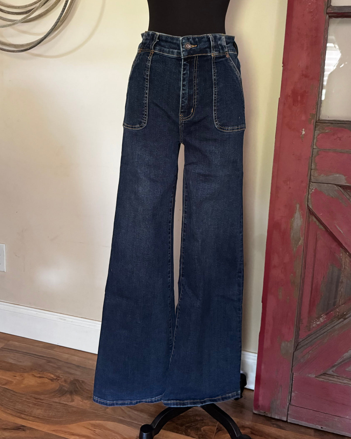 Dark blue wide-leg denim jeans displayed on a black mannequin torso in an indoor room with a beige wall, wood floor, and a distressed red door frame.